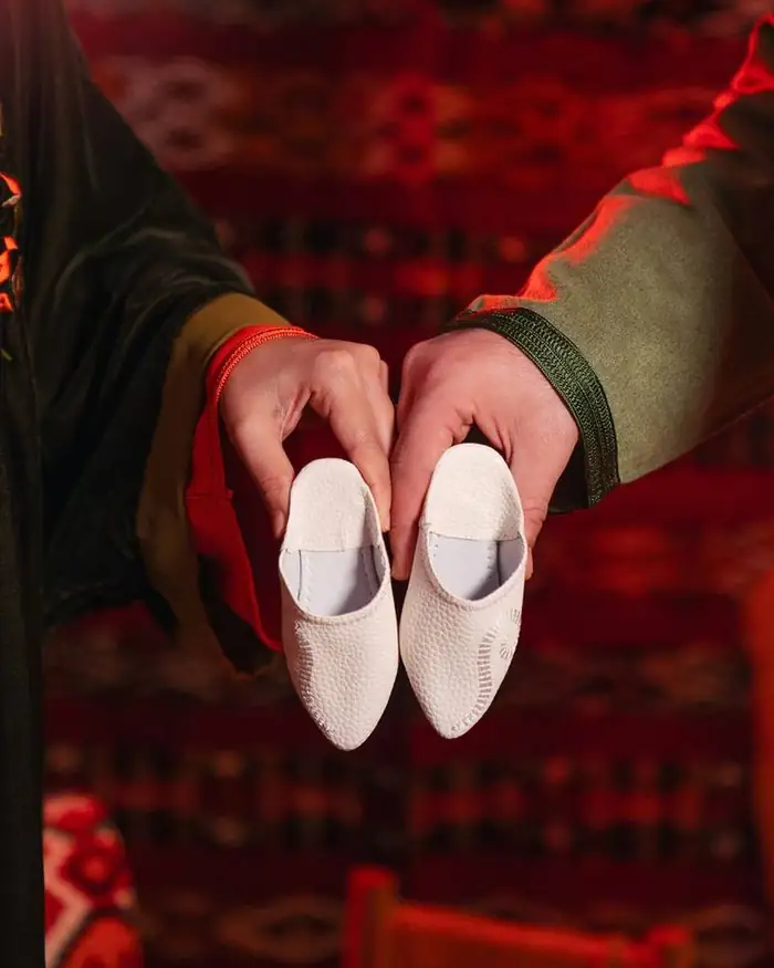 Two pairs of hands presenting white leather babouches over a red Moroccan rug